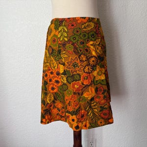 May include: A vintage knee-length skirt with a vibrant floral pattern in shades of orange, yellow, and green on a brown background. The skirt is displayed on a mannequin, showcasing its A-line shape and overall design.