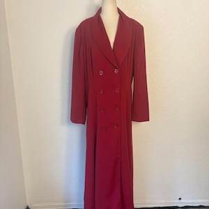 May include: A long, double-breasted, red dress with a notched collar and six matching buttons. The dress is displayed on a mannequin against a neutral wall. The dress has long sleeves and a floor-length hemline, suitable for formal occasions.