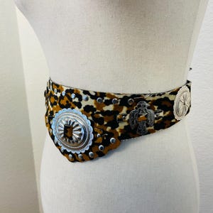 May include: A leopard print belt with silver-tone metal embellishments. The belt features a repeating pattern of brown and black spots on a cream background. Decorative silver-tone metal pieces include a large oval buckle, a bird, and small studs.