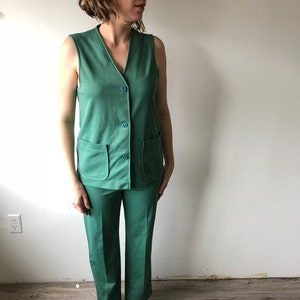 May include: A vintage green pantsuit featuring a sleeveless vest with two front pockets and three blue buttons. The matching pants have a straight leg design. The outfit is made of a solid green fabric.