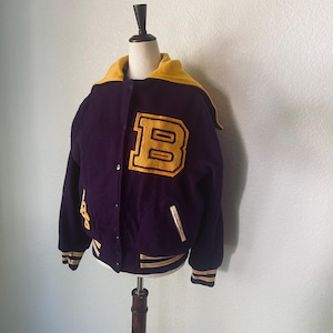 Op de afbeelding: Een paarse en gouden varsity-jas met een grote gouden "B" patch op de borst. De jas heeft een gele kraag en manchetten met gouden strepen. De jas is dichtgeknoopt en wordt op een mannequin getoond.