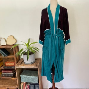 May include: A long, teal robe with a burgundy upper section and white trim. The robe has a matching belt and pockets. It is displayed on a mannequin. The robe is made of a soft, plush material.