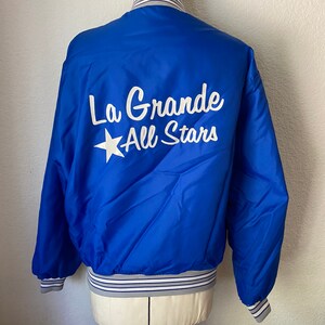 K&ouml;nnte beinhalten: Eine blaue Satin-Bomberjacke mit einem wei&szlig;en Stern und dem Text "La Grande All Stars" auf der R&uuml;ckseite. Die Jacke hat wei&szlig;e und graue gestreifte B&uuml;ndchen und einen Bund.