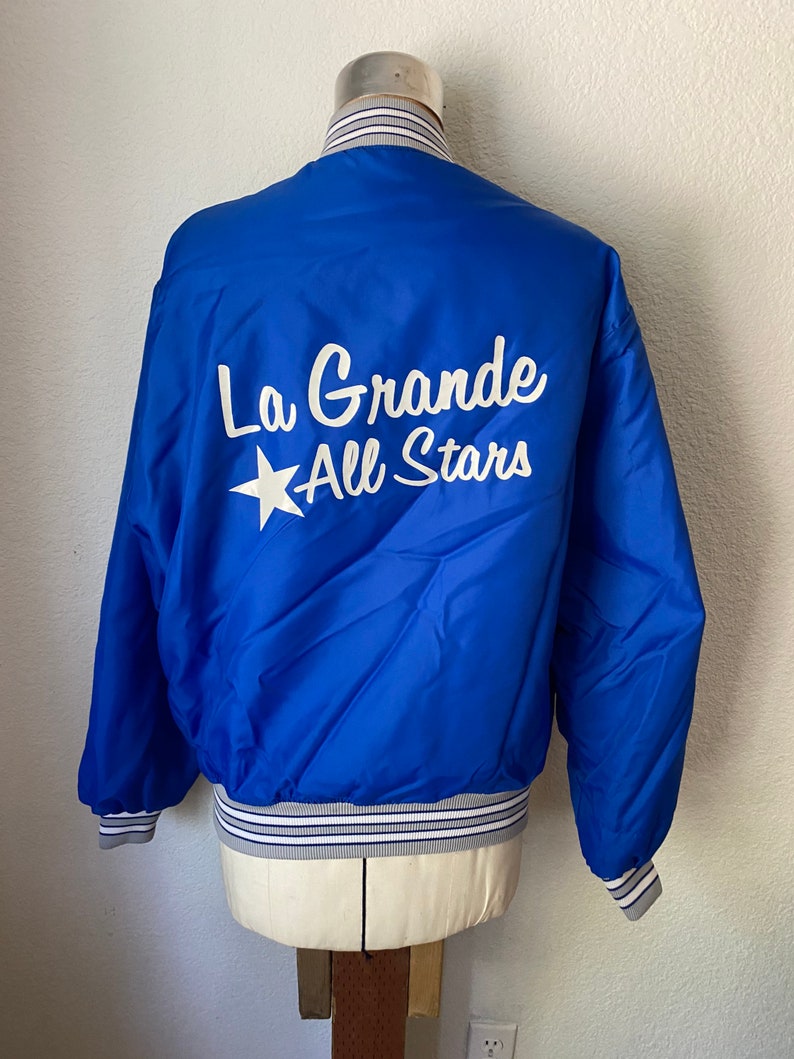 K&ouml;nnte beinhalten: Eine blaue Satin-Bomberjacke mit einem wei&szlig;en Stern und dem Text "La Grande All Stars" auf der R&uuml;ckseite. Die Jacke hat wei&szlig;e und graue gestreifte B&uuml;ndchen und einen Bund.