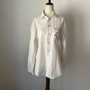 May include: White button-down shirt with long sleeves, displayed on a mannequin. The shirt features decorative embroidery along the button line and on the pocket, which has a colorful illustration. The shirt is a classic style.