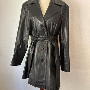 May include: A dark charcoal gray leather coat featuring a notched collar, long sleeves, and a front button closure. The coat includes a matching leather tie belt at the waist and two side pockets, displayed on a mannequin torso.
