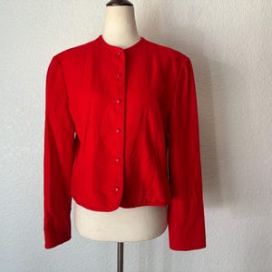 May include: A bright red, button-down jacket with a round collar. The long-sleeved jacket is displayed on a mannequin. The jacket is a solid color and appears to be made of a smooth fabric. The garment is a classic style.