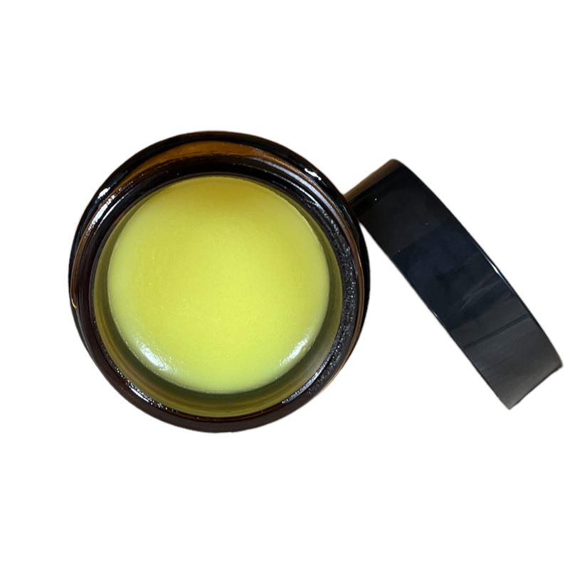 May include: A small glass jar with a yellow balm inside. The jar has a dark brown lid and a black lid.