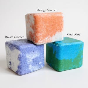 May include: Three colorful bath bombs in cube shapes. The top bath bomb is orange and white, labeled "Orange Soother." Below is a blue and white bath bomb labeled "Dream Catcher." The bottom right bath bomb is blue and green, labeled "Cool Aloe."