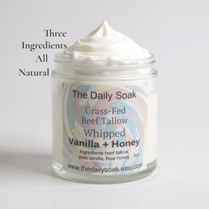 May include: A clear glass jar filled with a whipped, creamy product, topped with a swirl. The label reads "The Daily Soak" and lists ingredients: grass-fed beef tallow, vanilla, and honey. Text on the jar reads "Three Ingredients All Natural".