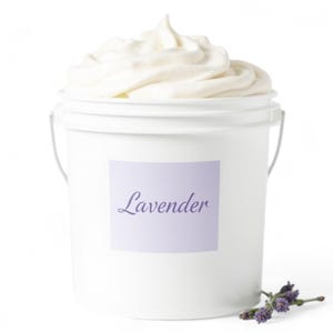 May include: A white bucket filled with a swirl of white cream, labeled "Lavender" on a light purple sticker. A sprig of lavender flowers rests near the bucket. The image is set against a white background.