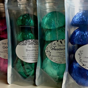 May include: Four clear plastic bags filled with shower steamers. Each bag contains several foil-wrapped, mushroom-shaped steamers in shades of pink, green, and blue. Each bag has a label with text.