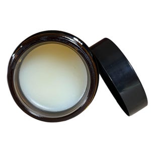 May include: A clear balm in a brown glass jar with a black lid. The jar is open and the balm is visible.