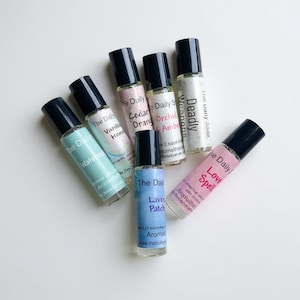 May include: An assortment of essential oil roll-on bottles with black caps. The bottles are labeled with names like "Love Spell," "Watermelon," and "Lavender Patch." The bottles are arranged on a white surface.