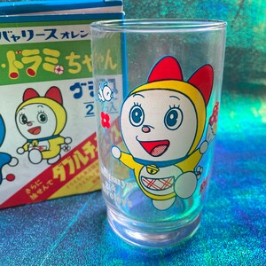 Puede incluir: Un vaso transparente con un personaje de dibujos animados con un atuendo amarillo, un lazo rojo y una campana. El personaje sostiene una flor y una seta. Una caja con texto japon&eacute;s est&aacute; en el fondo.