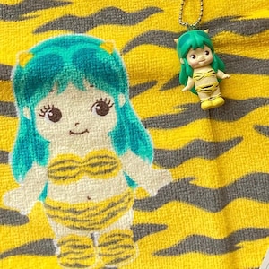 May include: A yellow and gray tiger-striped towel featuring a large embroidered graphic of a character with teal hair wearing a yellow and black tiger-striped bikini, accompanied by a small matching figurine keychain with a metal ball chain.