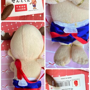 May include: A plush doll of a cartoon character with a light tan body, brown antlers, and blue shorts with red trim. The doll has gold accents and a red strap. A tag with Japanese text and the words "MADE IN CHINA" is attached.