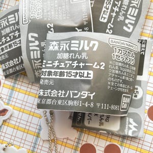 May include: Close-up of several small, gray and white packages with Japanese text. The packages are labeled with the words "Morinaga Milk" and "Miniature Charm 2". The packages are on a patterned surface with a few white charms.
