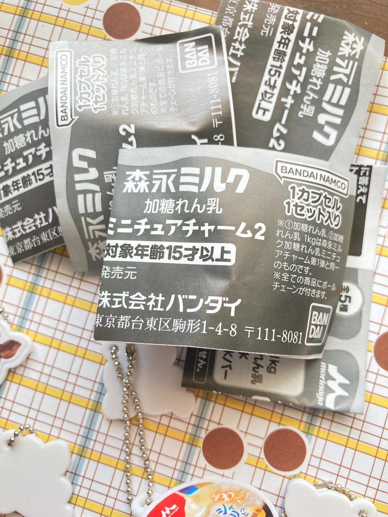 May include: Close-up of several small, gray and white packages with Japanese text. The packages are labeled with the words "Morinaga Milk" and "Miniature Charm 2". The packages are on a patterned surface with a few white charms.