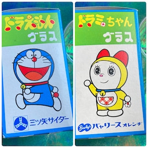 Puede incluir: Dos cajas de vidrio japonesas vintage con personajes de Doraemon. Una presenta un robot azul con forma de gato con collar y campana rojos. La otra caja muestra un personaje amarillo similar a Doraemon con capucha roja. Texto japon&eacute;s.