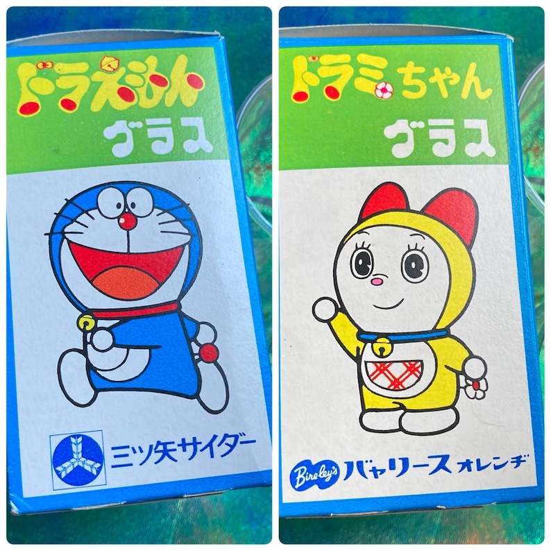 Puede incluir: Dos cajas de vidrio japonesas vintage con personajes de Doraemon. Una presenta un robot azul con forma de gato con collar y campana rojos. La otra caja muestra un personaje amarillo similar a Doraemon con capucha roja. Texto japon&eacute;s.