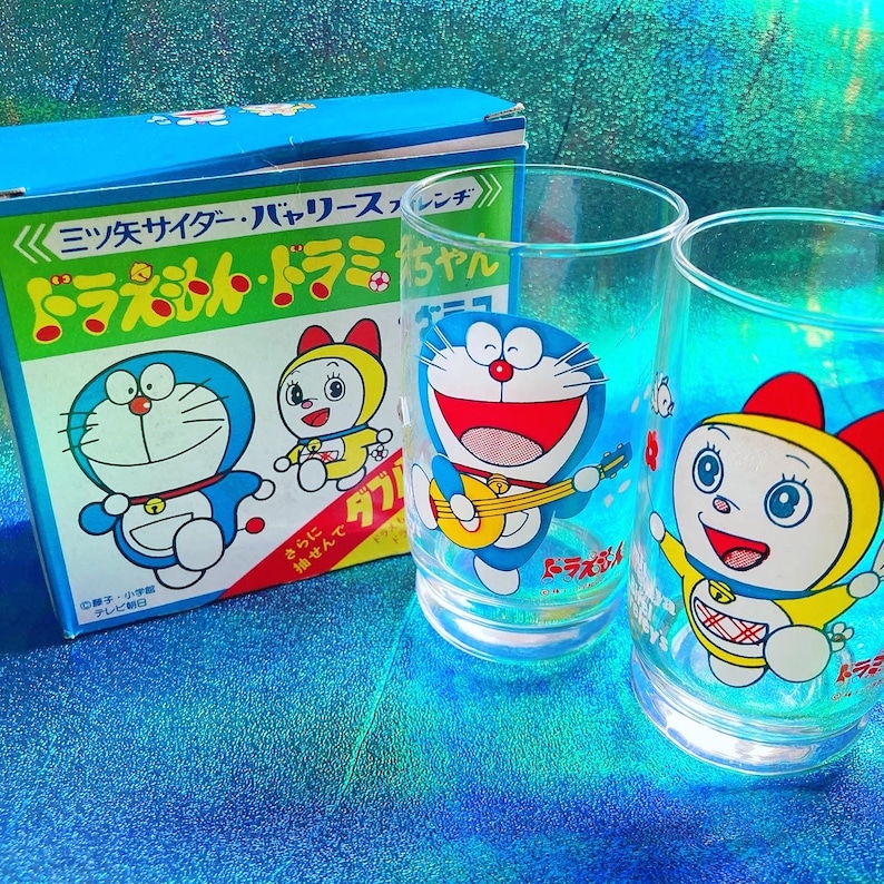 Puede incluir: Una caja azul y dos vasos transparentes con los personajes de dibujos animados Doraemon y Dorami. La caja tiene texto japon&eacute;s e ilustraciones de los personajes. Los vasos tienen dise&ntilde;os coloridos.