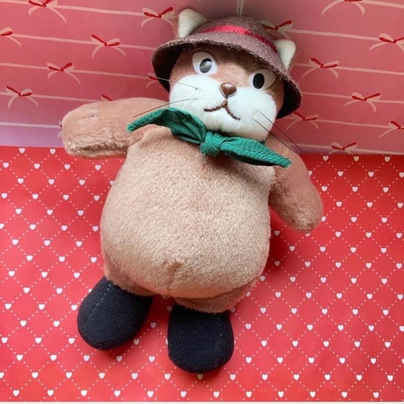 May include: A plush toy cat with a brown hat, green bow tie, and black shoes. The cat has a tan body and white face with whiskers. The background is a red surface with white hearts and bows.