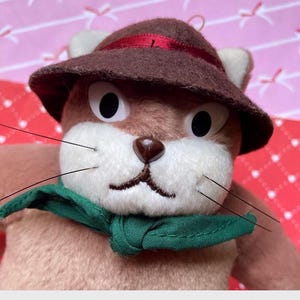 May include: A plush toy squirrel with a brown hat and a green scarf. The squirrel has a white face, black eyes, and long whiskers. The hat is brown with a red band. The toy is a light brown color.