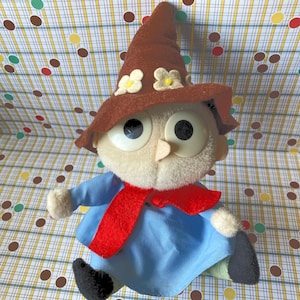 May include: A vintage plush doll with a whimsical design. The doll features a brown pointed hat adorned with white and yellow flower accents, large round eyes, and a red scarf. It wears a blue tunic and black shoes, evoking a storybook character.