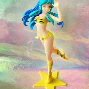 Puede incluir: Figura de un personaje de anime femenino con cabello azul y un bikini amarillo y negro. La figura está de pie sobre una base en forma de estrella amarilla, con un brazo levantado. La figura lleva botas amarillas.