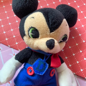 May include: A plush Mickey Mouse doll with black ears, a black face, and a tan muzzle. The doll wears a blue outfit with red buttons and a red bow tie. The doll has green embroidered eyes and white gloves.