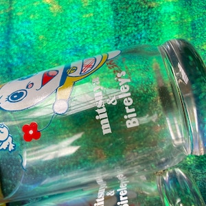 Puede incluir: Botella de vidrio transparente con un dise&ntilde;o de personaje de dibujos animados y el texto "mitsuya & Bireley's". La botella presenta un personaje de dibujos animados blanco con detalles en rojo, azul y amarillo. Tambi&eacute;n se ve una flor roja.