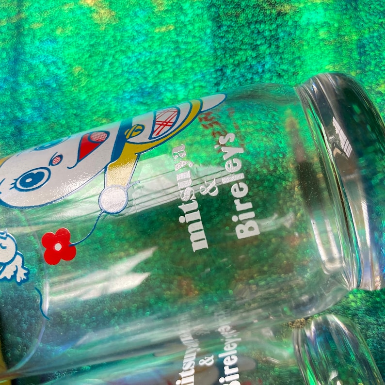 Puede incluir: Botella de vidrio transparente con un dise&ntilde;o de personaje de dibujos animados y el texto "mitsuya & Bireley's". La botella presenta un personaje de dibujos animados blanco con detalles en rojo, azul y amarillo. Tambi&eacute;n se ve una flor roja.