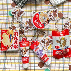 May include: A collection of Japanese-themed keychains featuring dairy products. The keychains include cow-shaped charms, ice cream, and condensed milk packaging. The color palette is primarily red, white, and brown, with Japanese text.
