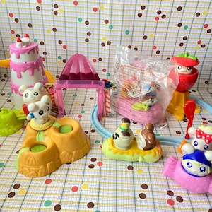 Op de afbeelding: Een verzameling Hamtaro-speelgoedfiguren van plastic, waaronder een roze taart met lagen, een paarse bergstructuur, een gele rotsbasis met twee figuren, een klein bootje met twee figuren, een rood appelvormig speeltje en een figuur in een plastic zak.