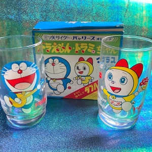 Puede incluir: Dos vasos transparentes con ilustraciones de personajes de dibujos animados. Un vaso presenta un personaje azul con forma de gato tocando la guitarra, y el otro tiene un personaje amarillo sosteniendo flores. Una caja azul con texto japon&eacute;s est&aacute; detr&aacute;s de los vasos.