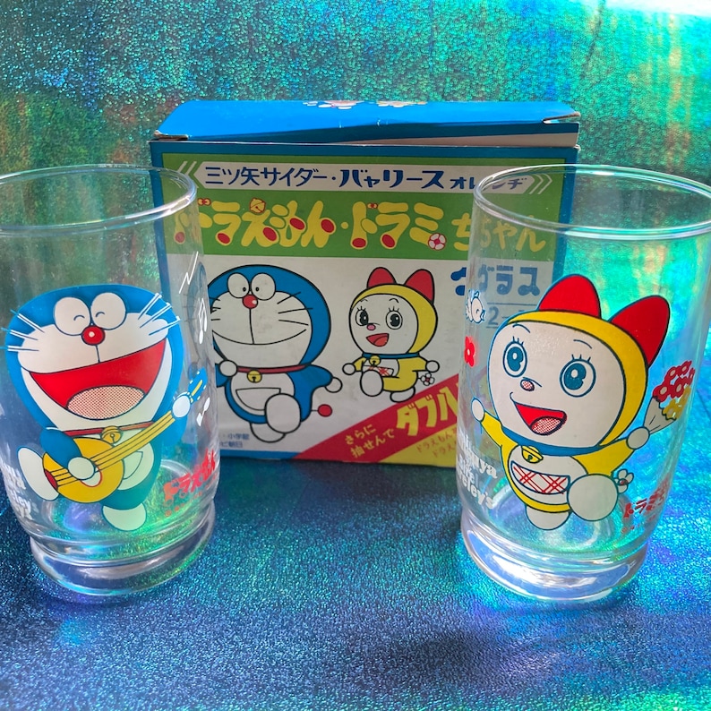 Puede incluir: Dos vasos transparentes con ilustraciones de personajes de dibujos animados. Un vaso presenta un personaje azul con forma de gato tocando la guitarra, y el otro tiene un personaje amarillo sosteniendo flores. Una caja azul con texto japon&eacute;s est&aacute; detr&aacute;s de los vasos.