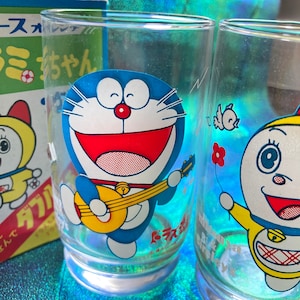 Puede incluir: Dos vasos transparentes con ilustraciones del personaje de dibujos animados Doraemon. Un vaso presenta a Doraemon tocando la guitarra, mientras que el otro tiene un personaje sosteniendo una flor. Una caja con texto japon&eacute;s es visible en el fondo.