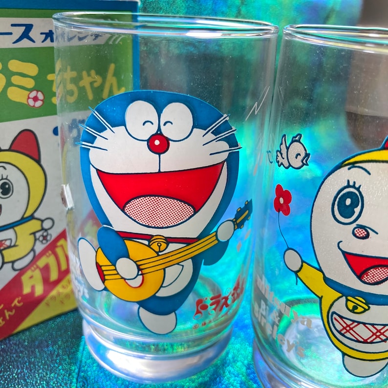 Puede incluir: Dos vasos transparentes con ilustraciones del personaje de dibujos animados Doraemon. Un vaso presenta a Doraemon tocando la guitarra, mientras que el otro tiene un personaje sosteniendo una flor. Una caja con texto japon&eacute;s es visible en el fondo.