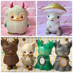 May include: A collection of six small vinyl figures featuring a white furry monster with pink horns, a white creature with a gold mushroom cap, two brown and white deer-like figures, and two green and gray figures holding small oval frames.