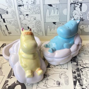 Puede incluir: Dos figuras de cerámica de hipopótamos de dibujos animados. Uno es amarillo con pelo naranja, el otro es azul claro. Ambos están reclinados sobre bases blancas en forma de nube. El fondo presenta una tira cómica.