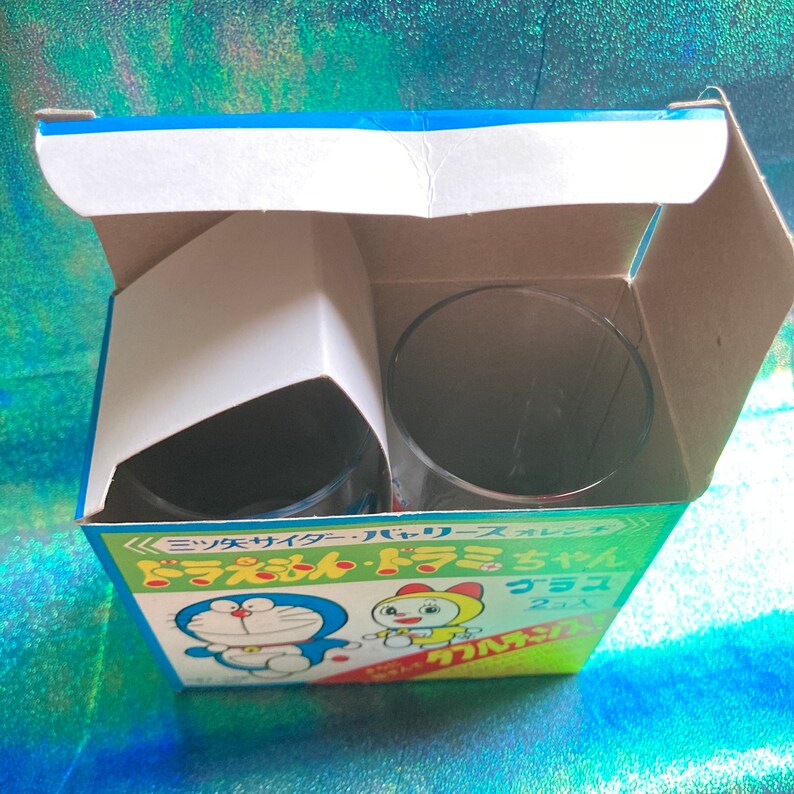 Puede incluir: Una caja con dos vasos transparentes. La caja es azul, verde y amarilla con texto japon&eacute;s y personajes de dibujos animados. Los vasos son visibles dentro de la caja abierta.