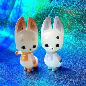 May include: Two adorable fox figurines with large ears and black eyes. One fox has gold accents on its ears and scarf, the other has silver accents. Both are wearing scarves and have a cute, cartoonish design.