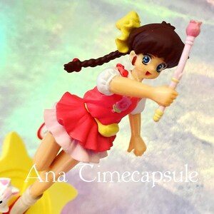 Puede incluir: Figura de una niña con un vestido rojo y una falda rosa, sosteniendo una varita rosa con una flor en la parte superior. Su cabello castaño está peinado con un lazo amarillo y una trenza larga. El texto "Ana Cimecapsule" es visible.