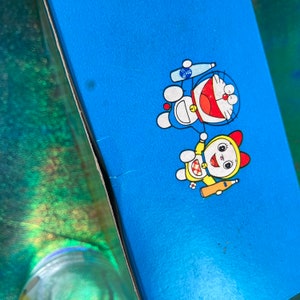 Puede incluir: Una caja azul con los personajes de dibujos animados Doraemon y Dorami. Doraemon es azul con un cuello rojo y una cara blanca, sosteniendo una botella. Dorami es amarilla con un lazo rojo y una cara blanca, tambi&eacute;n sosteniendo una botella.