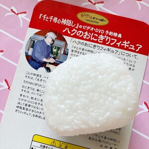 May include: A white, textured onigiri rice ball figurine, a promotional item for the movie "Spirited Away." The figurine rests on a red and white printed card with Japanese text and an image of a man.