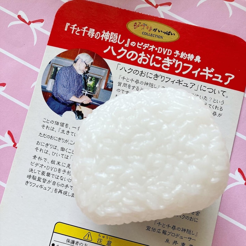 May include: A white, textured onigiri rice ball figurine, a promotional item for the movie "Spirited Away." The figurine rests on a red and white printed card with Japanese text and an image of a man.