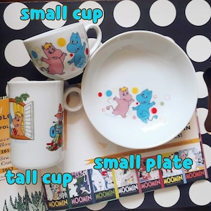 May include: A white ceramic small cup, tall cup, and small plate set. The small cup has a handle and features pink and blue cartoon characters. The tall cup has a handle and features a cartoon character. The plate has pink and blue cartoon characters.