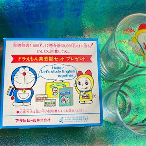 Puede incluir: Una caja azul con texto japon&eacute;s e ilustraciones de personajes de Doraemon, junto con dos vasos transparentes con dise&ntilde;os de Doraemon. La caja promociona un juego de conversaci&oacute;n en ingl&eacute;s.