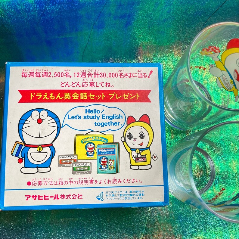 Puede incluir: Una caja azul con texto japon&eacute;s e ilustraciones de personajes de Doraemon, junto con dos vasos transparentes con dise&ntilde;os de Doraemon. La caja promociona un juego de conversaci&oacute;n en ingl&eacute;s.